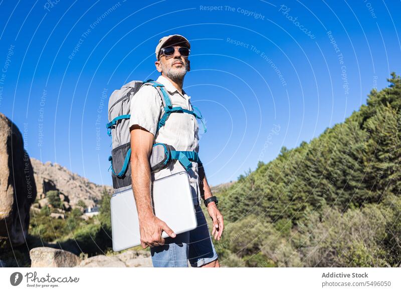 Man with laptop and backpack on cliff man hiker nature forest traveler rock summer journey male adventure rocky green gadget device environment woods activity