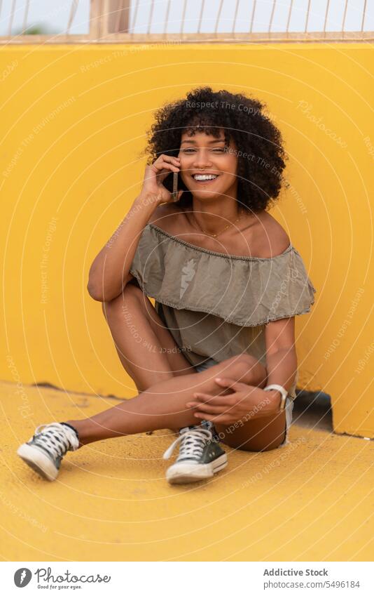 Happy woman talking via smartphone while sitting on yellow surface phone call cellphone using speak conversation smile young mobile african american communicate