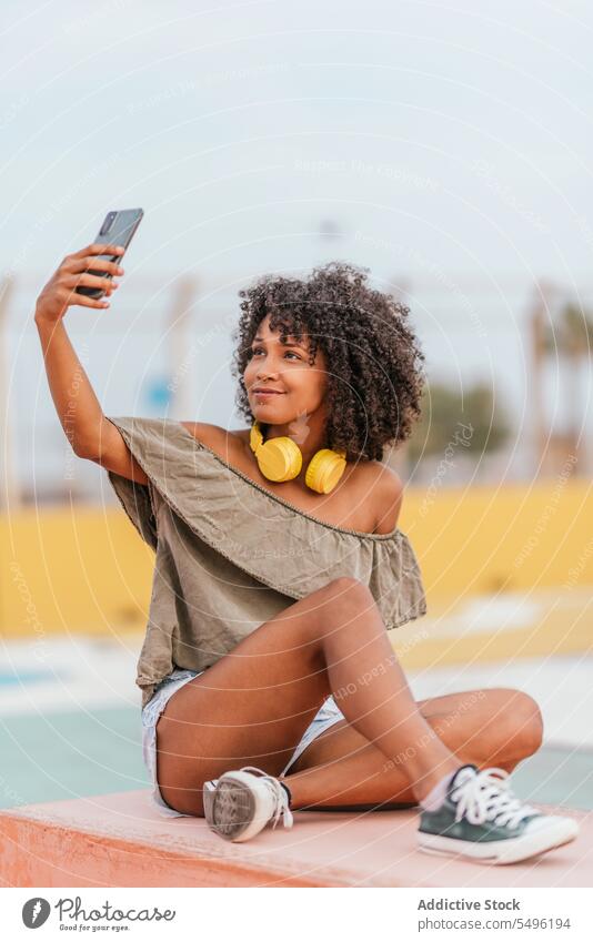 Smiling young female with headphones taking selfie on smartphone woman smile earphones using listen mobile happy positive bench device street enjoy cellphone