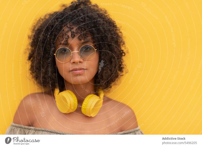 Young woman with headphones and eyeglasses looking at camera against yellow background piercing sunglasses curly hair model style modern trendy eyewear portrait