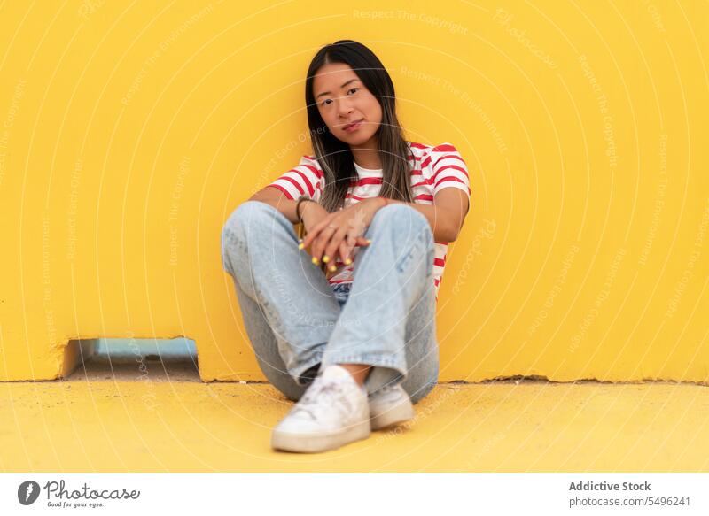 Asian woman sitting on floor against yellow background casual confident lean calm trendy outfit positive personality alone long hair lifestyle emotionless
