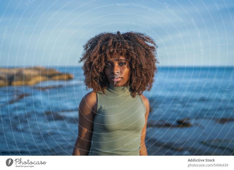 Serious African american young woman looking at camera while standing against blurred beach black lady female serious pensive thoughtful curly hair afro