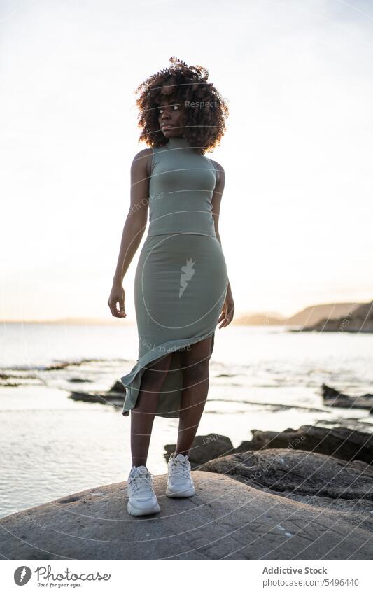 Serious African american young woman looking away while standing against blurred beach at sunset black lady female serious pensive thoughtful curly hair afro