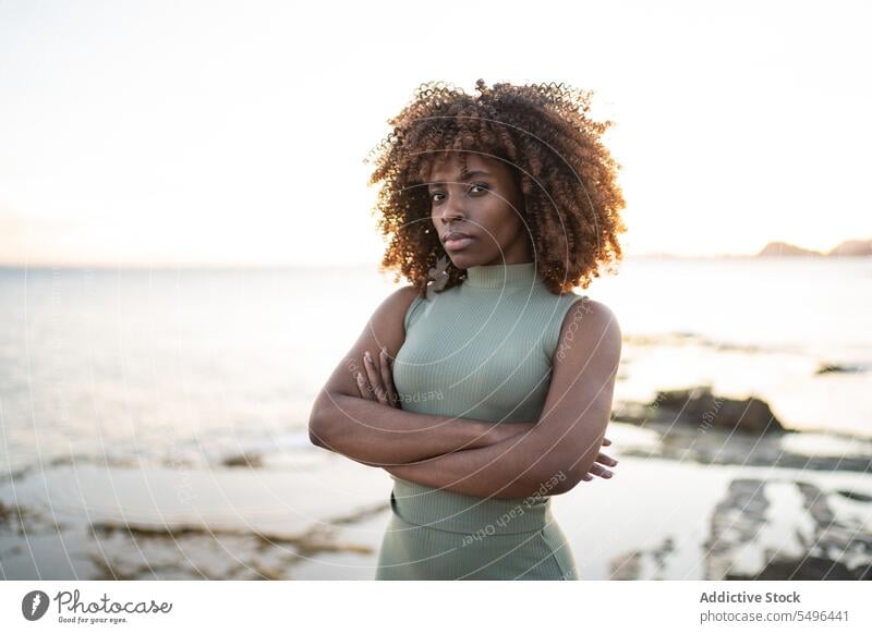 Serious African american young woman in dress looking at camera while standing against blurred beach at sunset black lady female curly hair afro