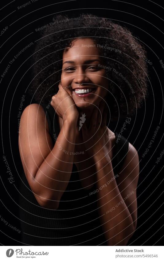 Cheerful ethnic woman touching cheek in dark studio positive touch face cheerful smile portrait appearance leisure carefree happy afro curly hair friendly young