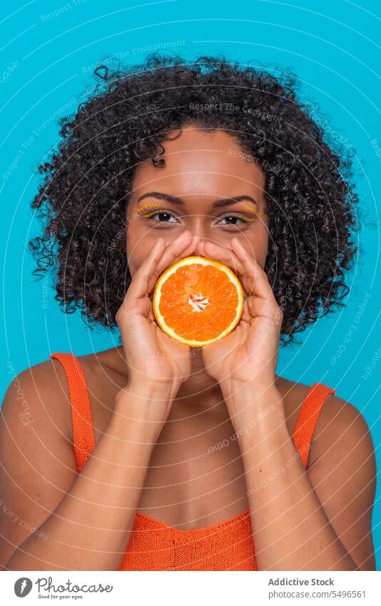 Happy ethnic woman covering mouth with orange slice smile happy citrus fruit skin care portrait young curly hair female hispanic positive bright joy model