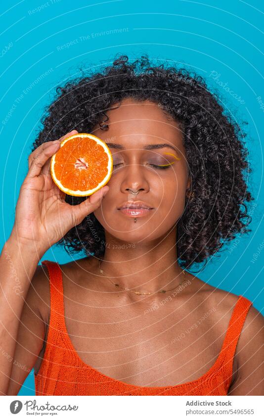 Serious ethnic woman covering eye with orange slice citrus fruit cover eye skin care portrait young curly hair female hispanic bright model appearance
