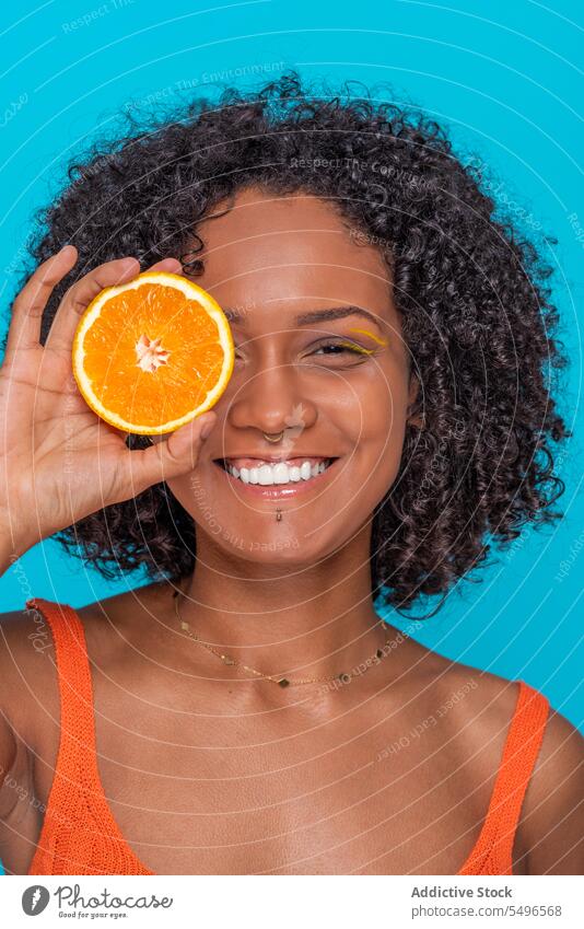 Smiling ethnic woman covering eye with orange slice smile happy citrus fruit cover eye skin care portrait young curly hair female cheerful hispanic positive