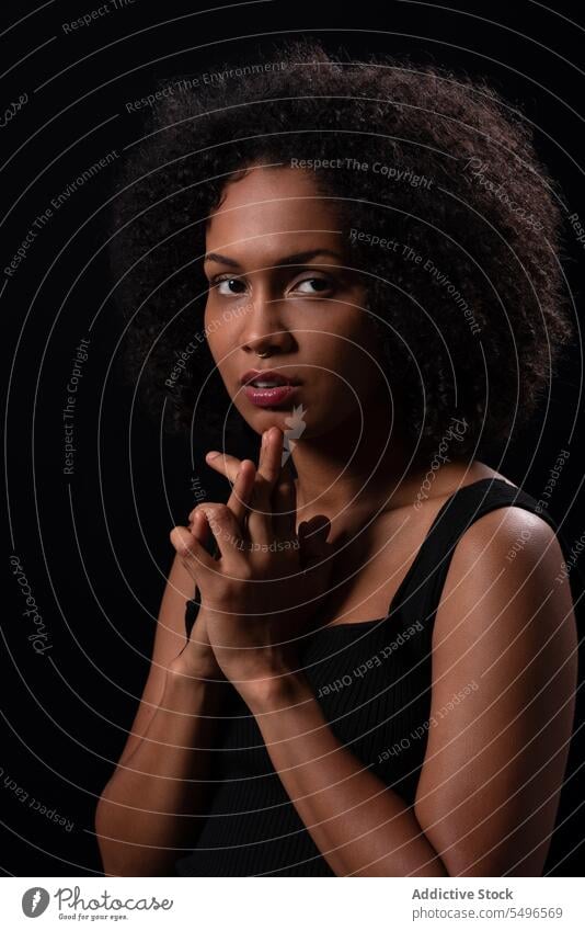 Thoughtful ethnic woman looking at camera in dark studio serious portrait hands clasped model individuality millennial pensive stare curly hair calm confident