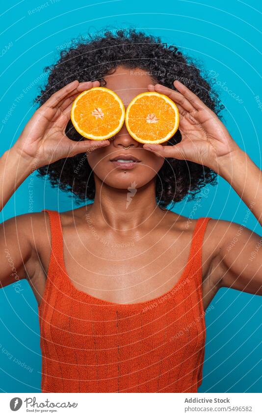 Serious ethnic woman covering eyes with orange slices in blue studio citrus fruit cover eye skin care portrait young curly hair female hispanic bright model