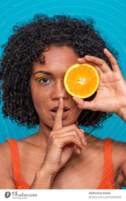 Woman covering eye with orange slice and gesturing silence woman cover eye portrait gesture silent fruit secret skin care citrus bright young curly hair female