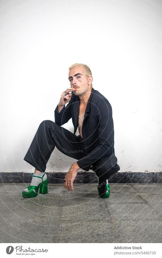 Serious man in high heels sitting in haunches and smoking on marble floor in daylight confident wall appearance posture serious individuality calm personality