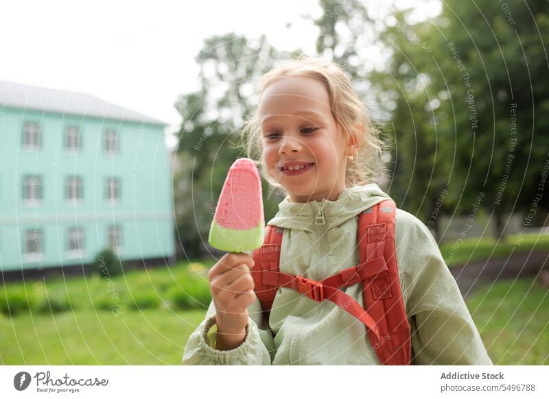 Positive kid with backpack standing in green park and enjoying ice cream in daylight child childhood grass happy eat cute adorable girl field carefree tasty
