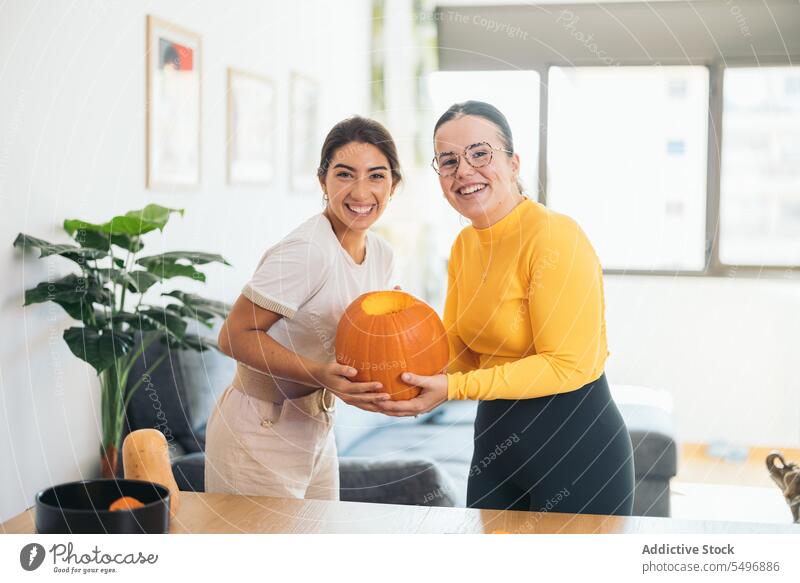 Happy ethnic young women standing with orange pumpkin in hands at home friend smile happy cheerful positive celebrate table female hispanic eyeglasses stripe
