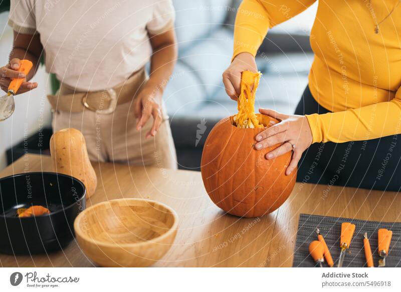Anonymous young women standing with orange pumpkin at home woman halloween pulp pull table female hispanic ethnic tool finger bowl utensil season work tradition