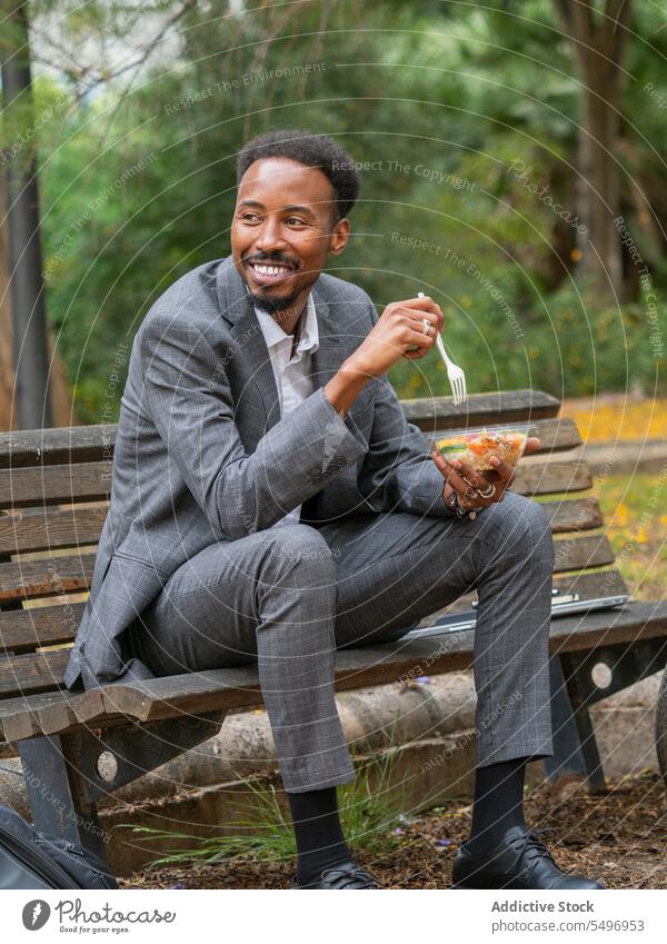 Happy black businessman sitting on bench in daylight and eating salad in bowl lunch fork smile healthy food happy park male young african american ethnic tasty