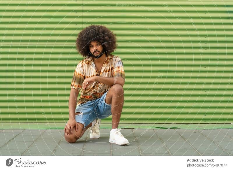 Stylish ethnic man with Afro hair against striped wall trendy style confident afro cool curly hair independent individuality crouch outfit appearance male young