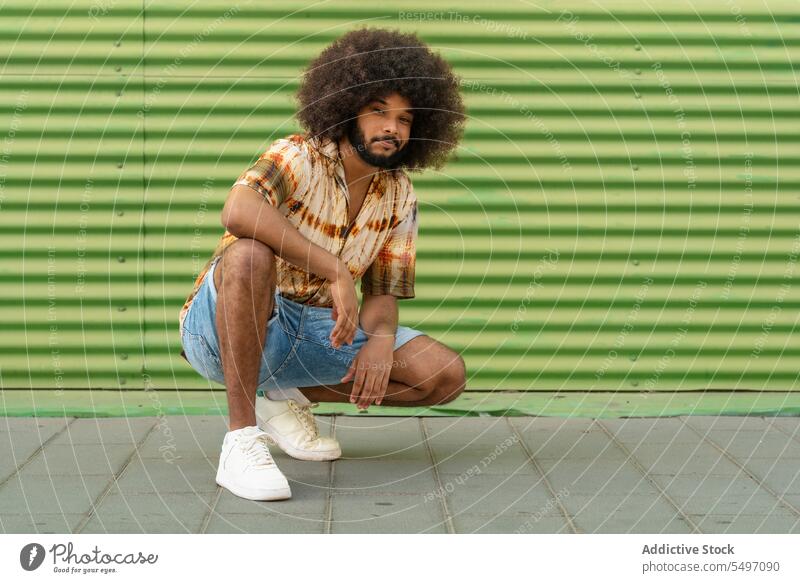 Stylish ethnic man with Afro hair against striped wall trendy style confident afro cool curly hair independent individuality crouch outfit appearance male young