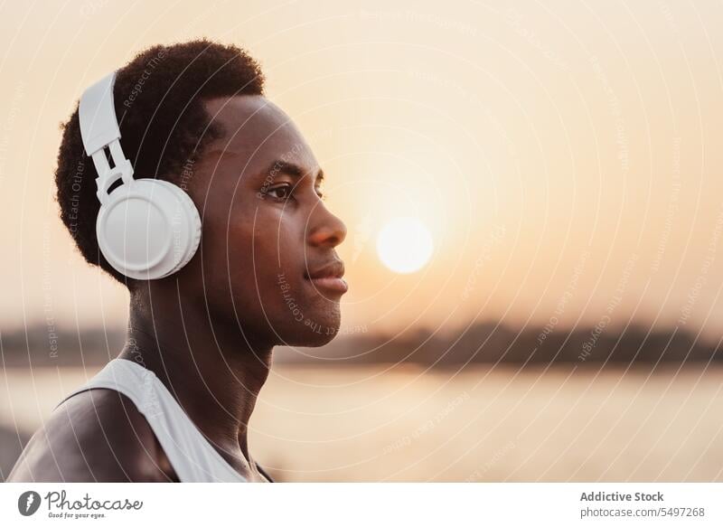 Black man listening to music in headphones near water sunset serious wireless song device male playlist gadget african american sundown evening modern coast
