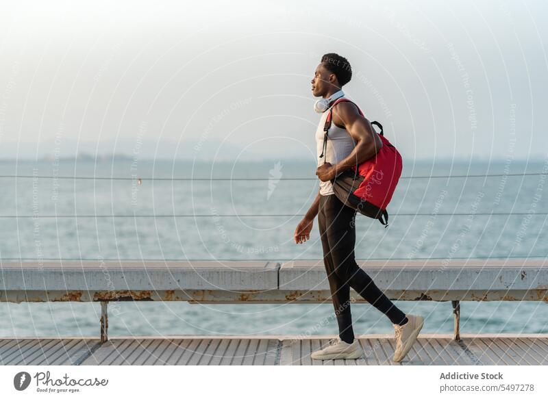 Black man with backpack and headphones near sea embankment walk water bag waterfront device seafront male african american black modern seaside fence ocean