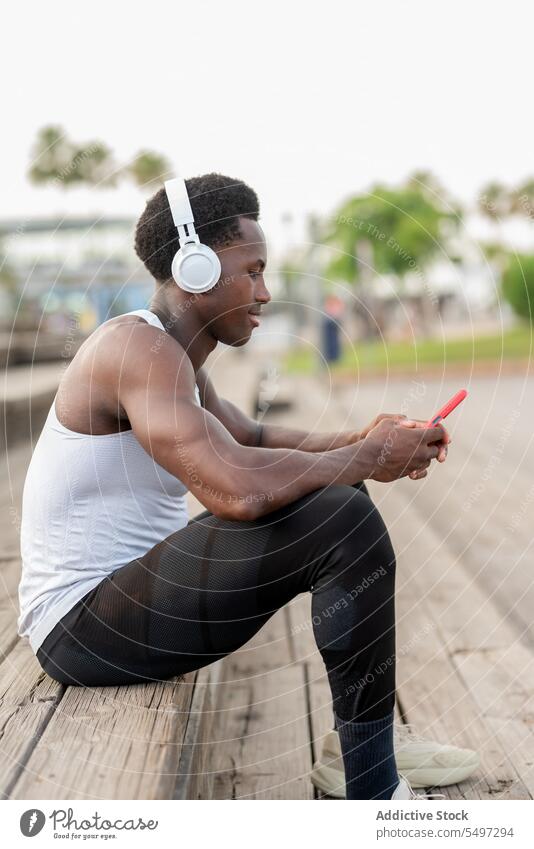 Delighted black sportsman in headphones with smartphone enjoying music athlete listen song text message staircase portrait male african american model fit