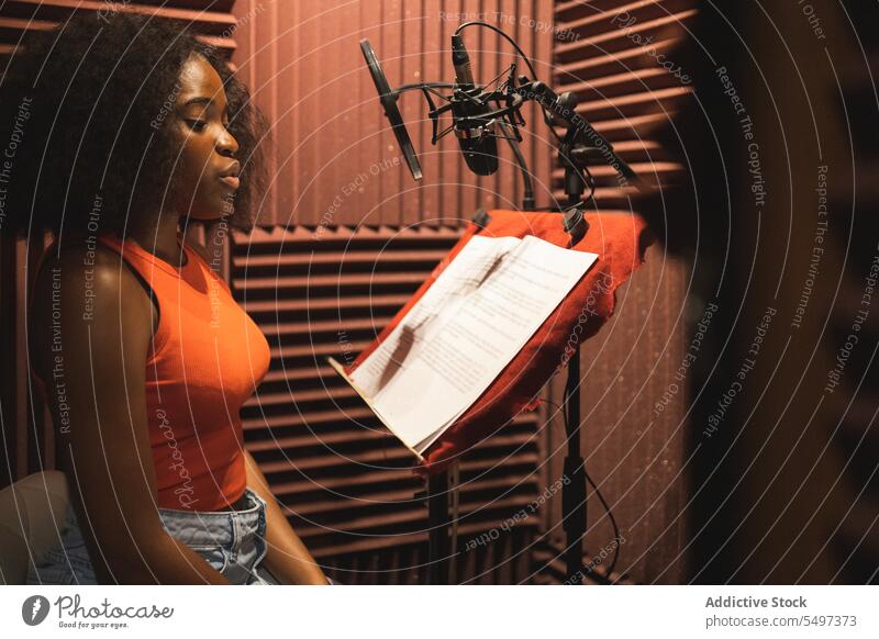 Focused black female standing and recording song near microphone equipment in studio woman singer paper focus audio young african american ethnic read sound