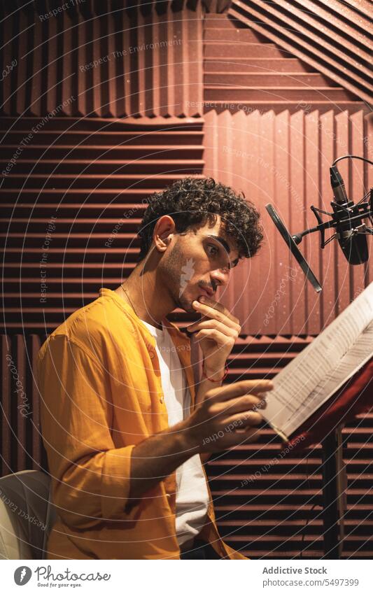 Thoughtful young ethnic man sitting and reading papers on desk in studio microphone serious hand at chin thoughtful pensive touch chin male hispanic curly hair