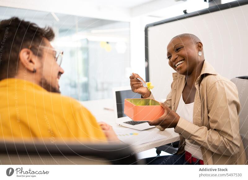 Cheerful African American woman sitting at a table with a friend and having lunch in the office colleague smile box happy break food cheerful female young black