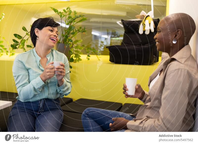Cheerful multiracial colleagues resting on cushion chairs in room with coffee cups women coffee break talk chat communicate smile conversation discuss cheerful