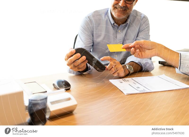 Smiling crop adult man at table with POS machine and unrecognizable female giving credit card woman smile shop payment purchase transaction customer manicure