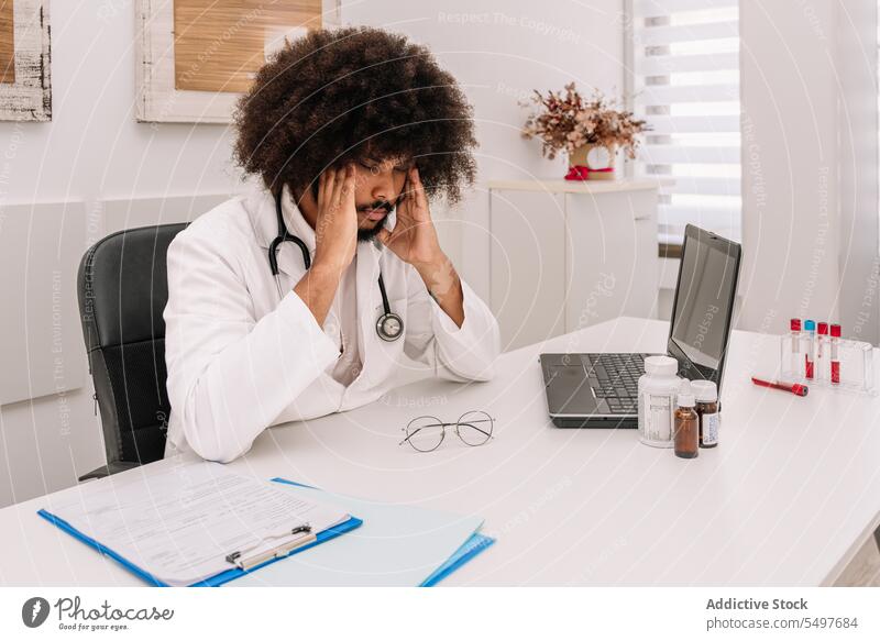 Thoughtful black doctor sitting at desk in hospital man stethoscope medical clinic pensive thoughtful lean on hand professional male job physician serious