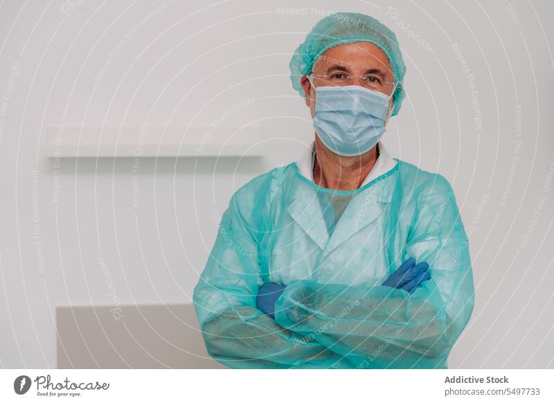Content male surgeon standing in clinic with folded arms man doctor arms crossed happy medic health care portrait hospital uniform professional mature mask
