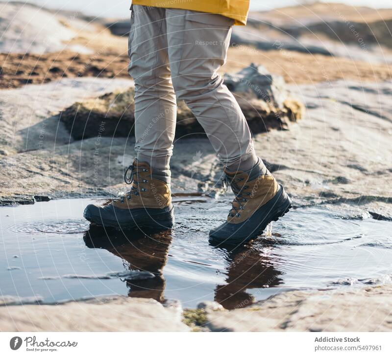 Crop person walking on puddle boot stone footwear rough water ground wet environment reflection weather natural feet sunlight shadow shade aqua rocky liquid