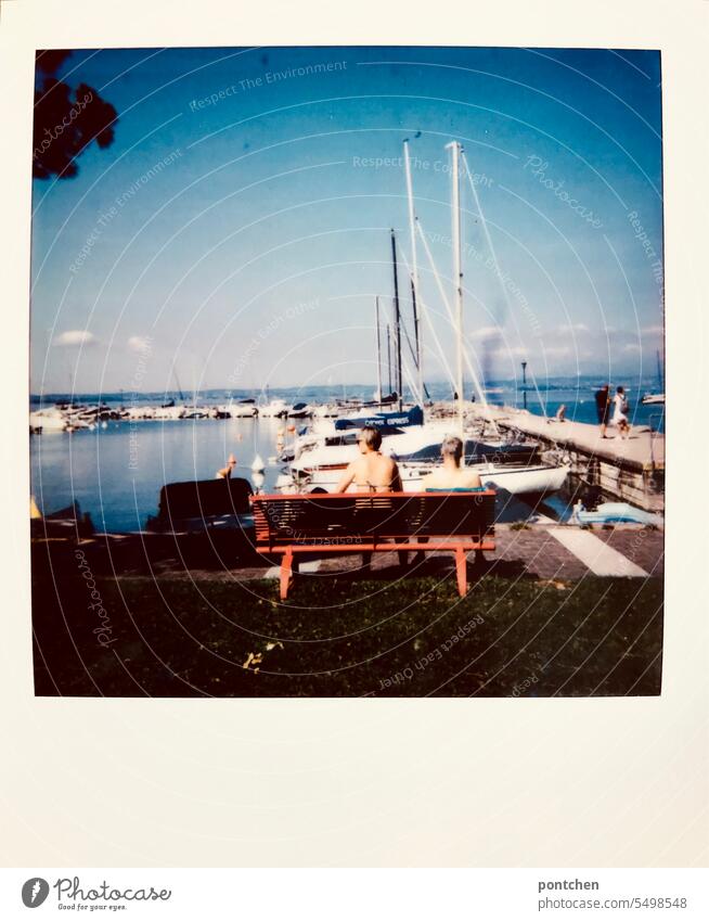 a couple in swimwear sits on a red bench at the harbor. lake garda Polaroid Couple Bench Lake Garda outlook Harbour Red senior citizens Back boats Exterior shot