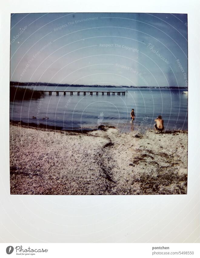 a father on the lakeshore in a crouch takes a picture of his son standing in the water. Polaroid Lakeside Beautiful weather Summer Lake Garda Sky