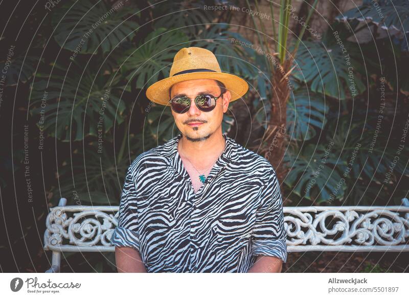 Middle-aged man with hat and sunglasses sits in shirt with zebra pattern on bench in front of large monstera plant Summer Man middle-aged man Hat Sunglasses