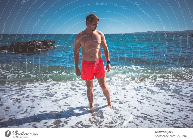 Middle-aged man with bright red swimming trunks stands in the sea and looks to the side Summer Summer vacation Summer's day go swimming Ocean Seashore Sea water