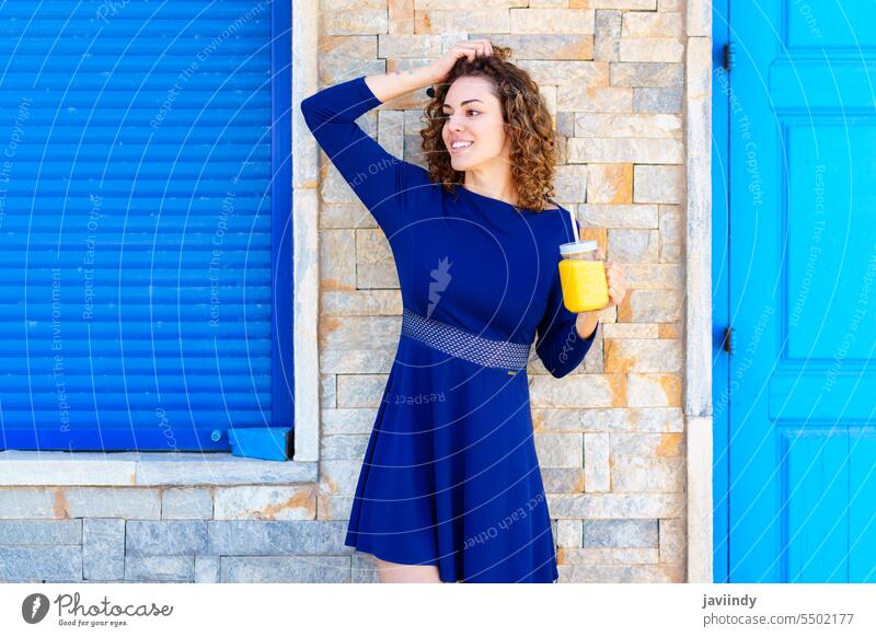 Smiling woman with juice on city street beverage smile touch head drink glass summer building shabby female positive young happy curly hair cheerful style fresh