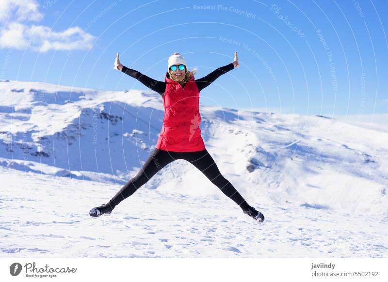 Cheerful woman in warm clothes jumping in air on snow terrain excited happy mountain winter active energy enjoy young female carefree freedom cheerful blue sky