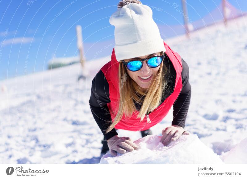 Smiling young woman in beanie wool cap and sunglasses warming up on snow lean on push up rock smile fit exercise happy positive female blond glove vest enjoy