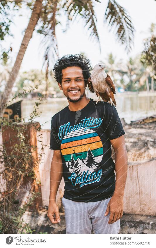 an eagle sits on a young mens shoulder, in the back are some palm trees Eagle male happy smiling person Bird outside outdoors portrait cheerful handsome smile