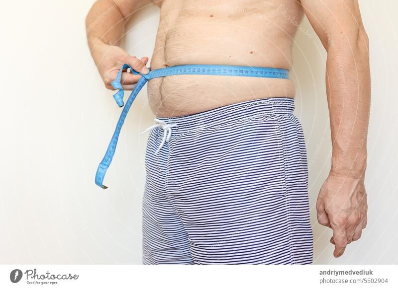 A man measures his fat belly with measuring tape. Concept of weight loss, health problems of obese people. Poor nutrition and sedentary lifestyle. Increased risk of heart illnesses. World Obesity Day