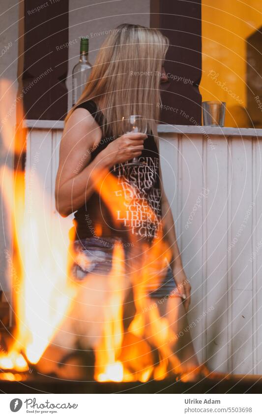 Drinkje bej Inkje | party - woman with long blond hair stands with wine glass in hand, behind campfire Warm-heartedness naturally Congenial kind Exterior shot