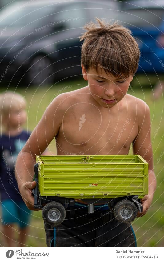 10 year old boy is carrying a trailer from a toy tractor. He is concentrated and looking forward to playing with his friends. In the background a smaller boy who is not recognizable and a green meadow with parked blurred cars.
