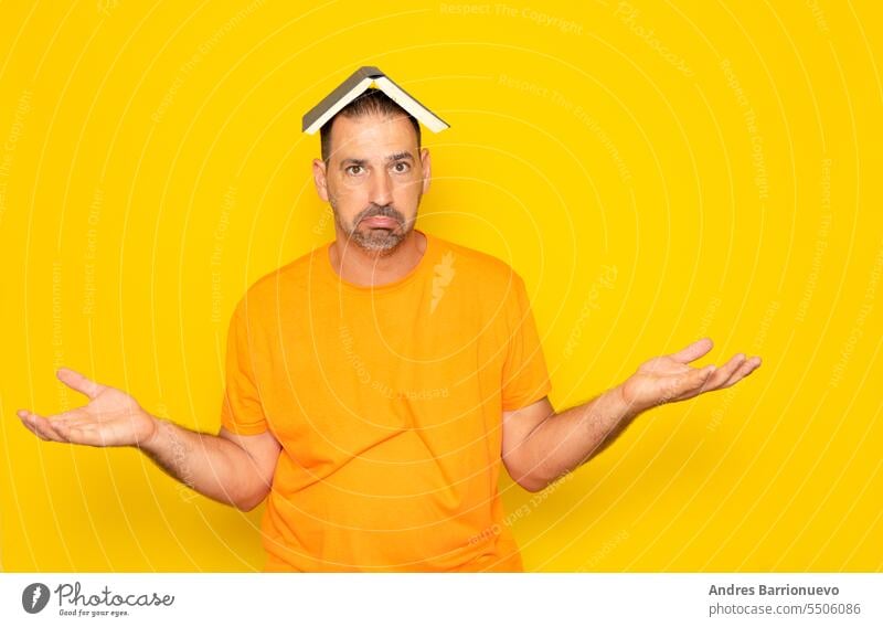 Bearded Hispanic man in his 40s wearing an orange t-shirt holding an open book on his head and raising his hands as he doesn't understand anything, isolated on yellow studio background