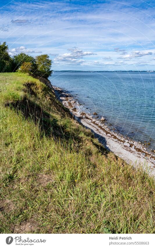 At the edge of the cliff, here it is overgrown with grass later also with trees. Below, the beach and the Baltic Sea run parallel to the cliff. The bright blue sky is drawn by white clouds