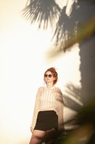 young beautiful red haired woman in casual clothes  in sunglasses with hair bun on beige wall background with palm tree shadows. Candid outdoor portrait in the park.