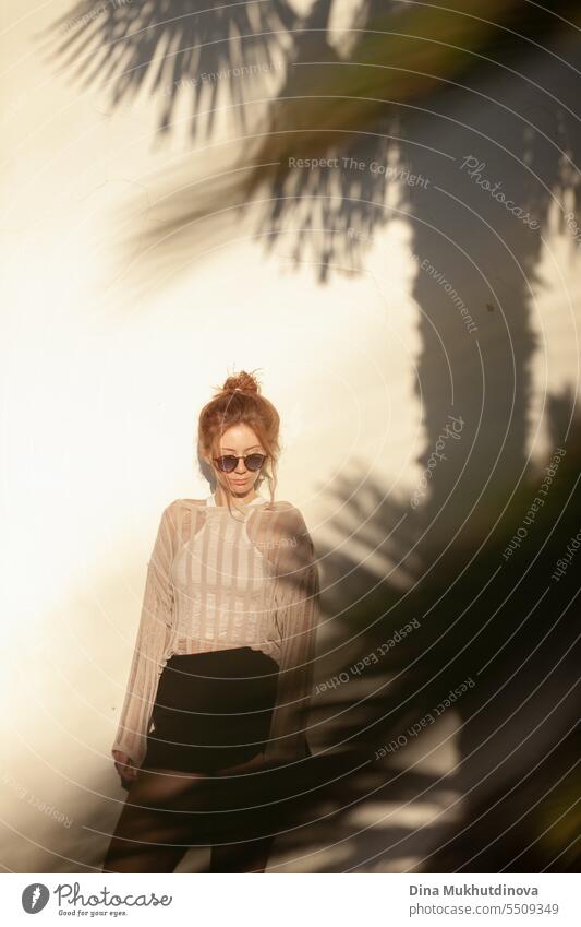 young beautiful red haired woman in casual clothes  in sunglasses with hair bun on beige wall background with palm tree shadows. Candid outdoor portrait in the park.
