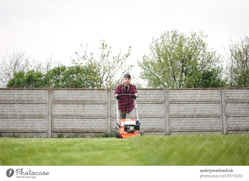 view of man in casual clothes mows lawn with lawn mower at backyard of his house. Husband takes care of garden on spring cloudy day. Modern gasoline garden equipment. Landscaping work