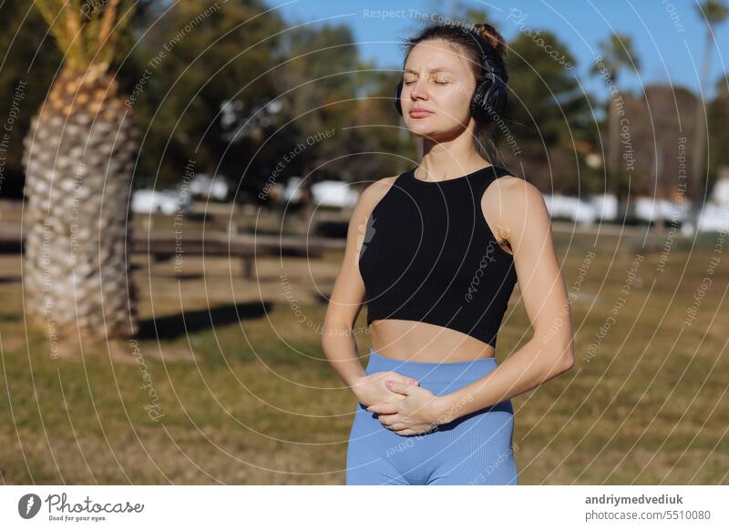 Concentrated young woman wearing headphones doing yoga meditation with online tutorial outdoors at park with palm trees. Listening music, harmony, calming, spirituality concept. World health day